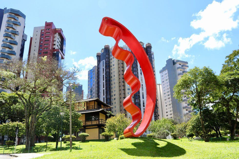 A red metal sculpture on a grassed plaza with a yellow Japanese building on the background.