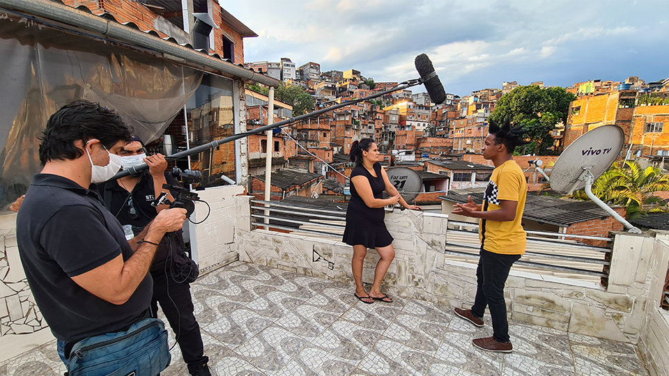 A man and a woman talk while a crew films  with the slums in the background