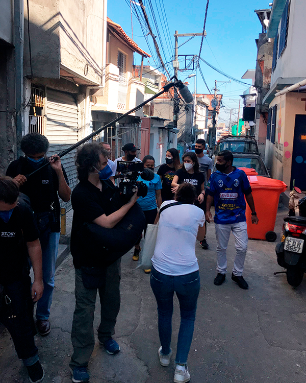 Tabata Amaral caminha em um beco estreito na favela com uma equipe filmando