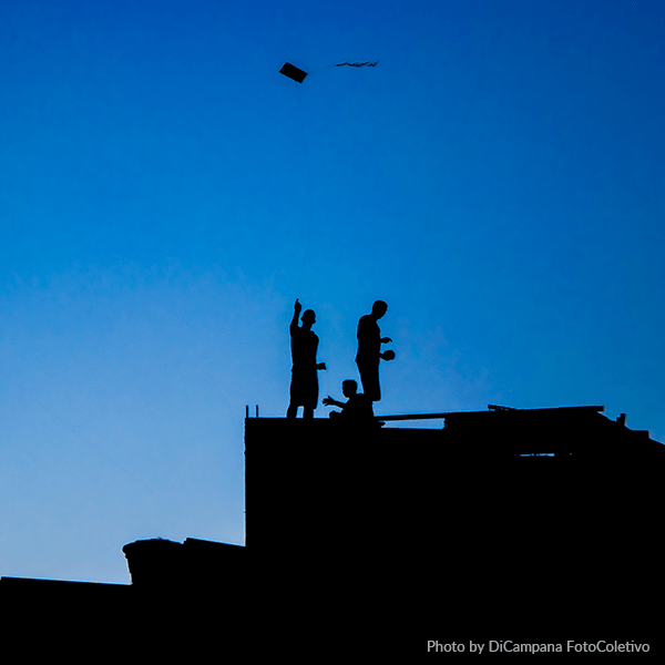 The shadows of 3 men on a rooftop flying kites agains a clear blue sky