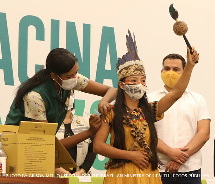 A masked indigenous woman holds a rattle high while being vaccinated by an indigenous masked nurse