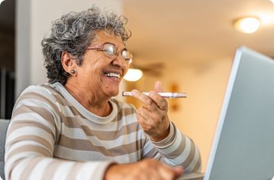 Elderly woman with glasses smiles, holding pen and using a laptop indoors.