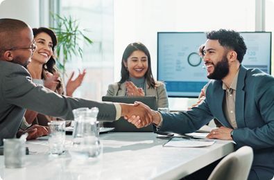 Two men shaking hands at a table in an office, surrounded by colleagues, some clapping.