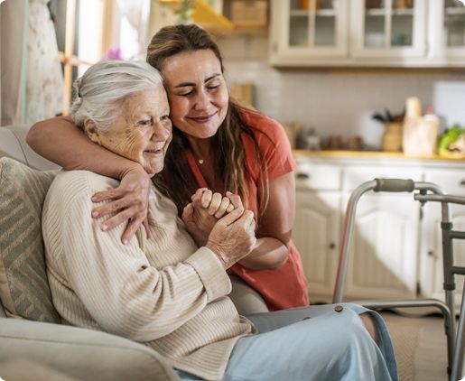 Woman hugging elderly woman, holding hands, smiles in kitchen, walker nearby.