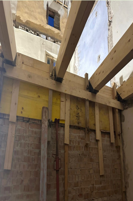 Interno parzialmente costruito con travi in legno a vista, muri in mattoni e un cielo nuvoloso visibile attraverso l'apertura sul tetto.