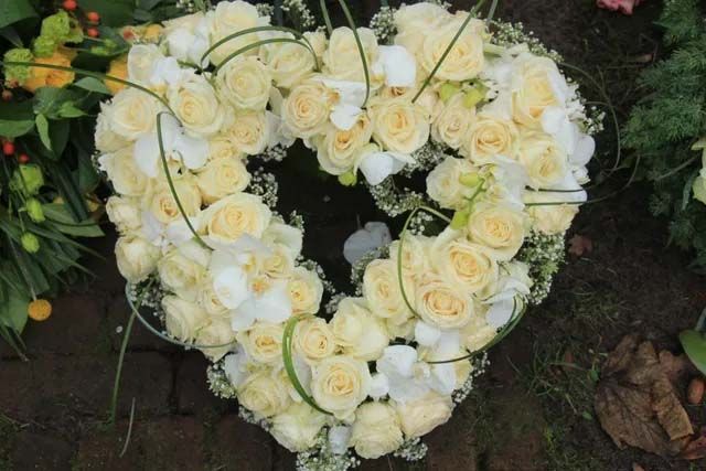 A heart-shaped funeral wreath made of light yellow roses and white orchids arranged on a green base.