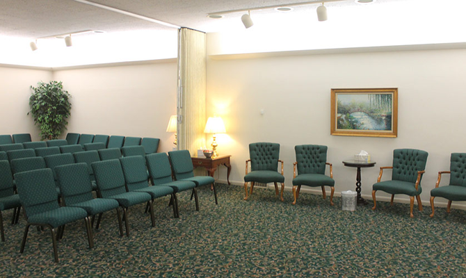 An aisle between white pews in a chapel with wooden floors, white walls, and ornate chandeliers overhead.