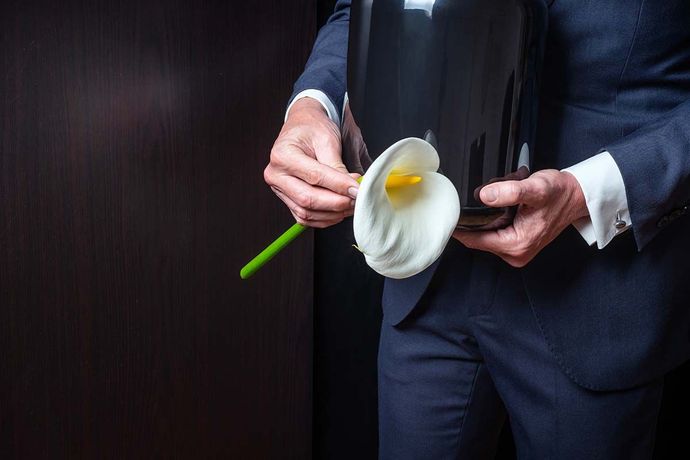 A person in a dark suit holds a black funeral urn and a single white calla lily against a dark background.