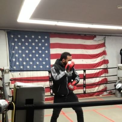 A man wearing boxing gloves stands in a boxing ring in front of an american flag