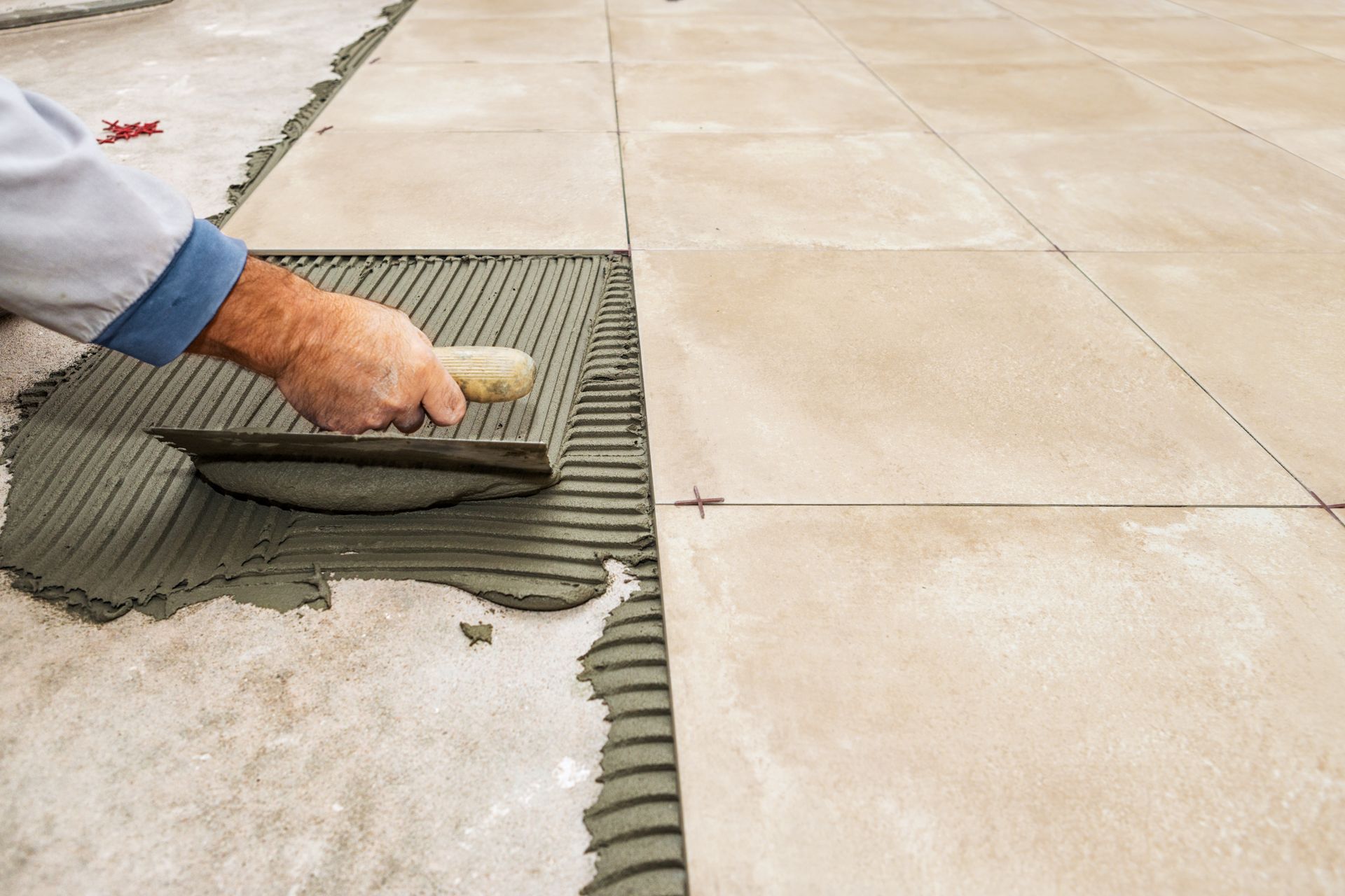 Worker spreading gray mortar on floor tiles beside installed beige tiles