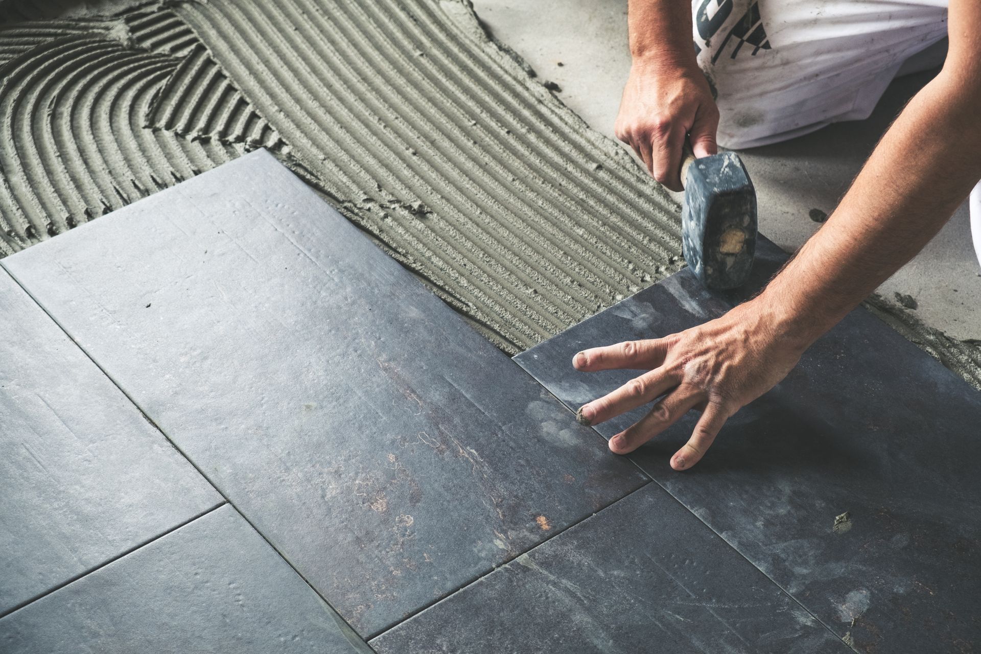 Hands using a mallet to install large gray floor tiles on a subfloor.