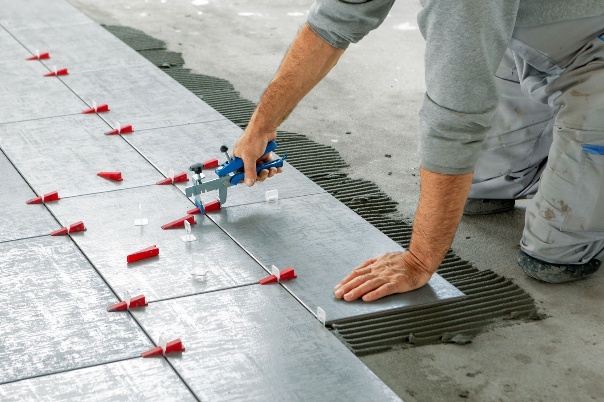 Worker leveling large gray floor tiles with red spacers during installation