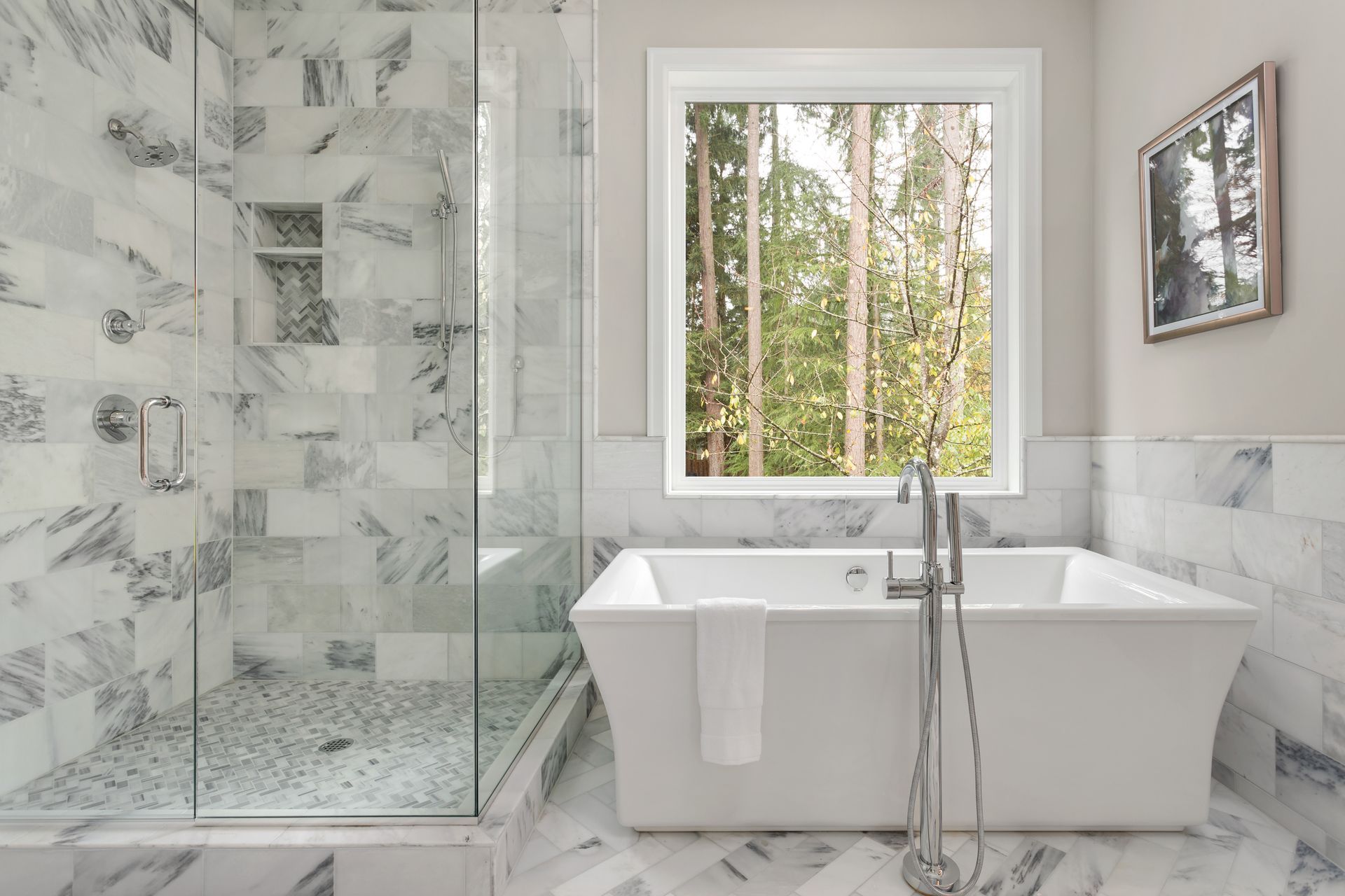 Modern marble bathroom with glass shower, freestanding tub, and window view of trees