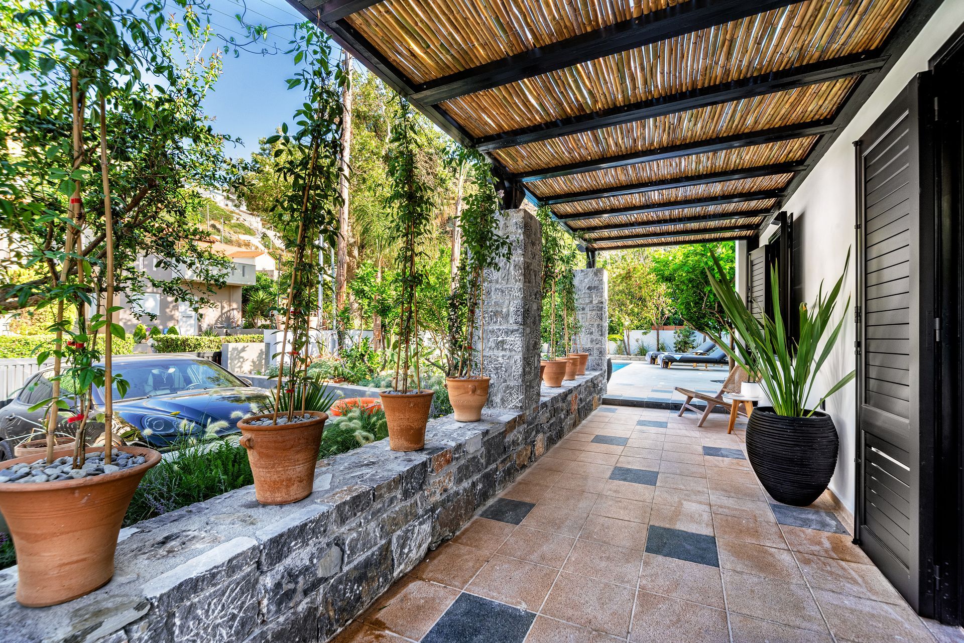 Covered stone patio with potted plants, wooden pergola, and black doors beside a garden view