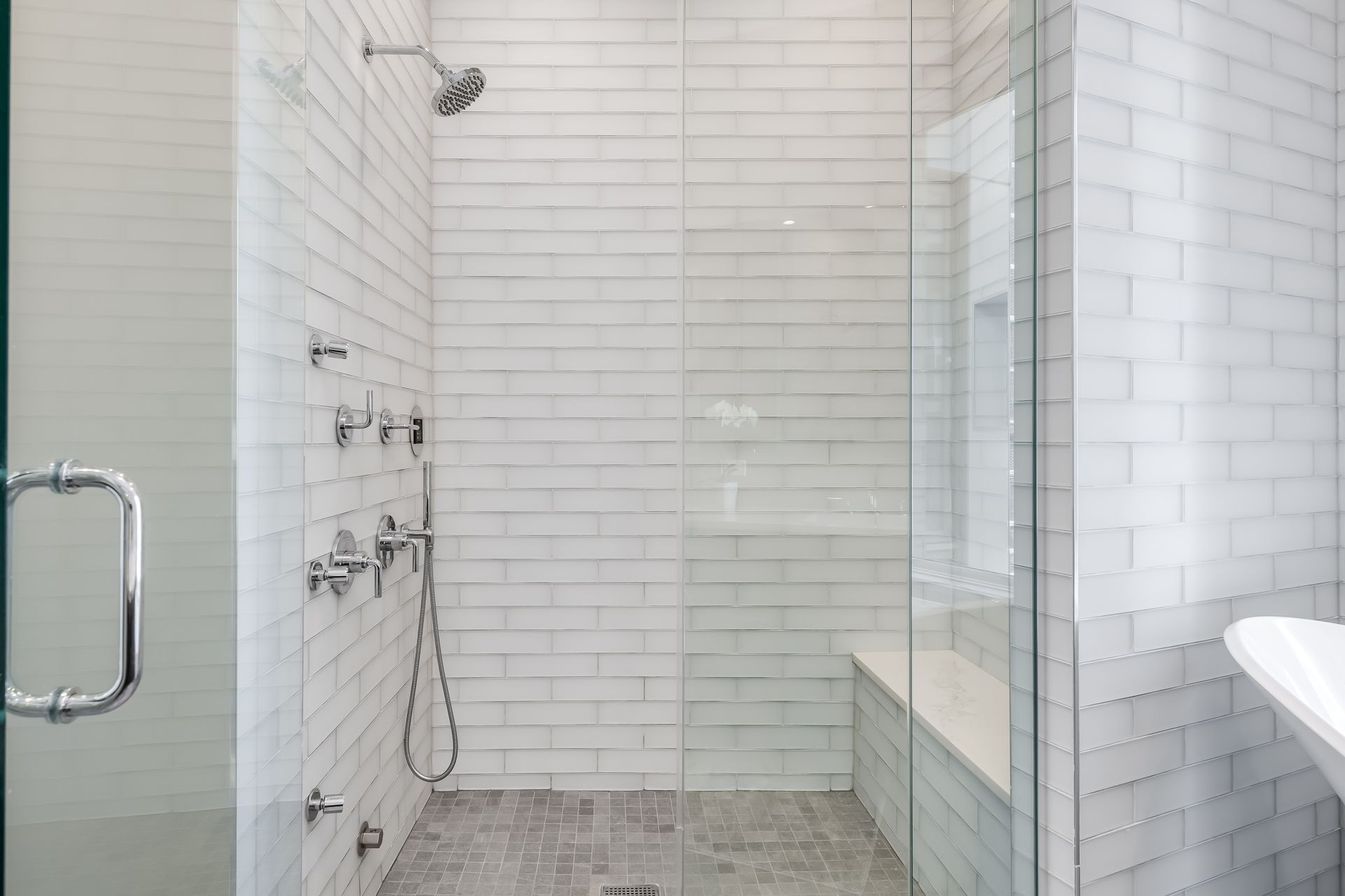 White tiled walk-in shower with multiple showerheads, a glass partition, and a metal grab bar