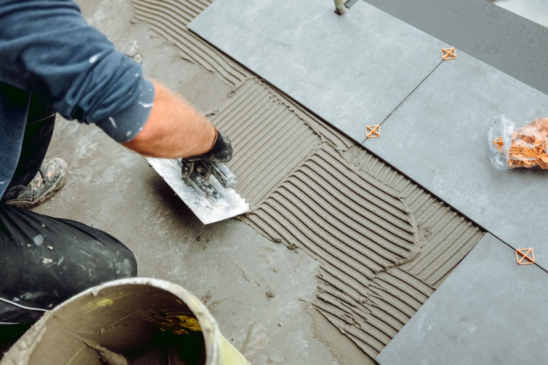Worker spreading gray tile adhesive on a floor with a trowel beside laid tiles