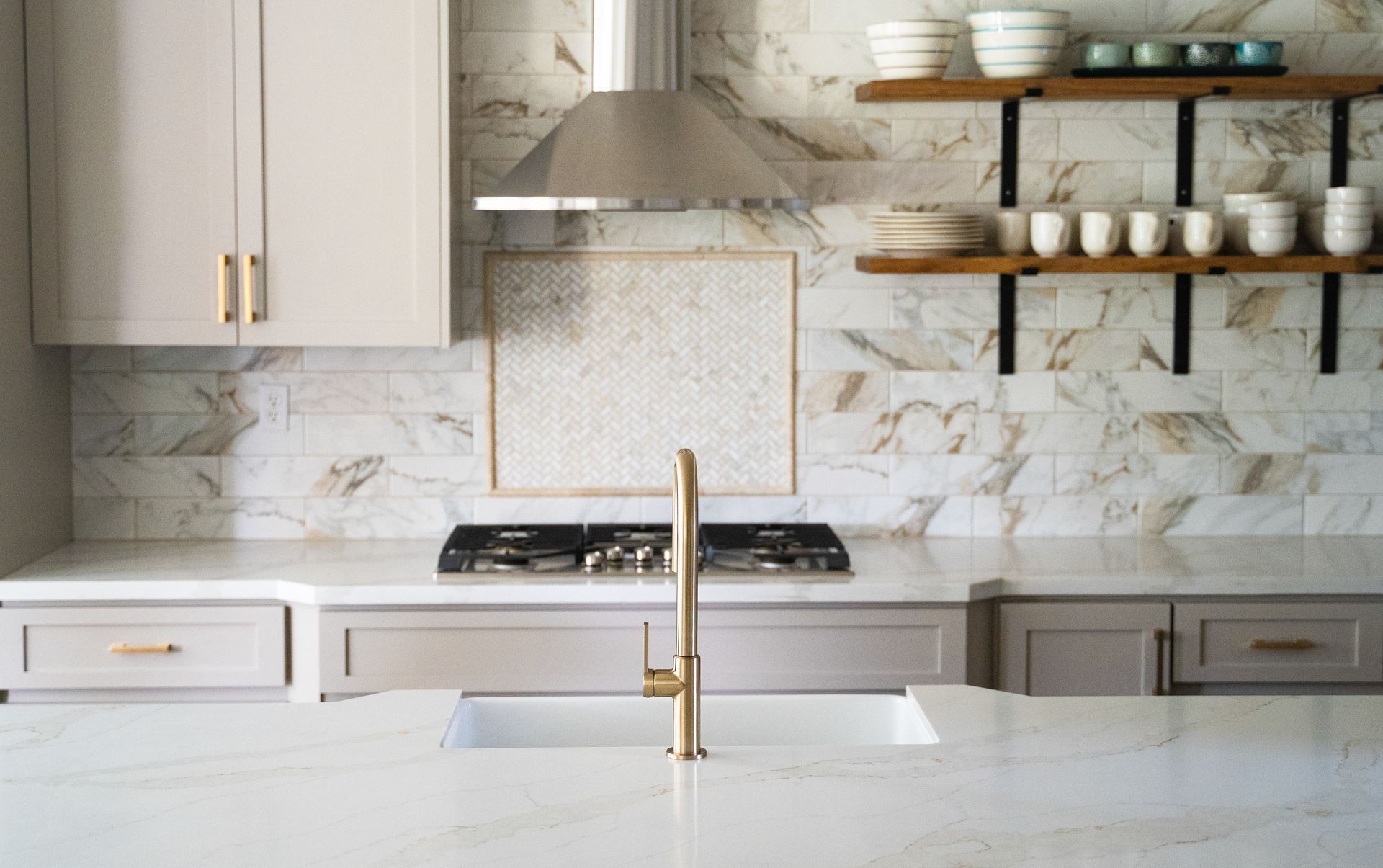 Modern kitchen with marble backsplash, white cabinets, open shelves, and gold fixtures around a central sink.