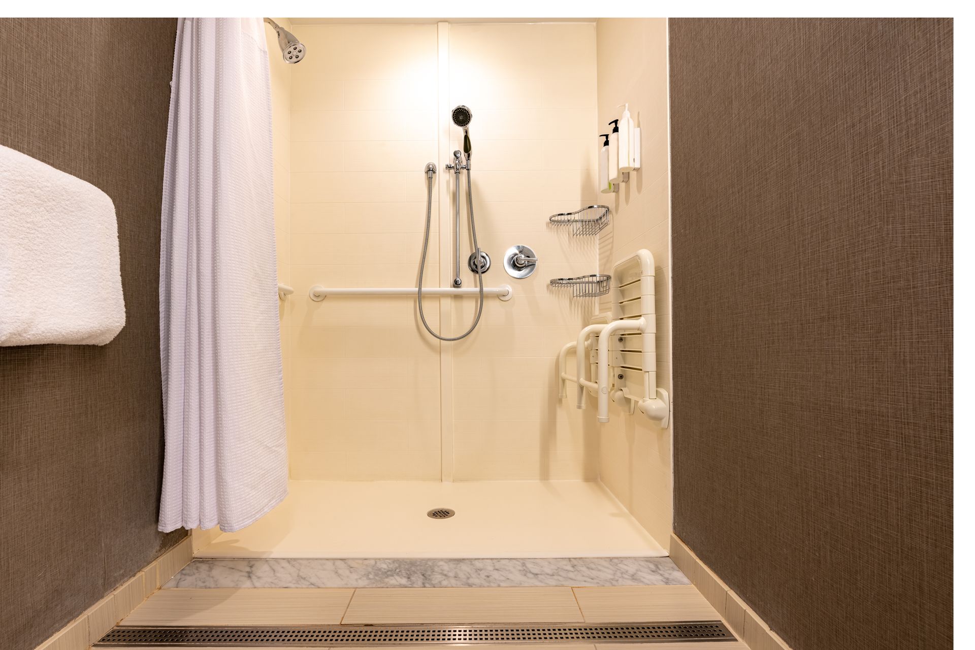 Small beige hotel shower stall with handheld showerhead, grab bars, and a curtain on the left