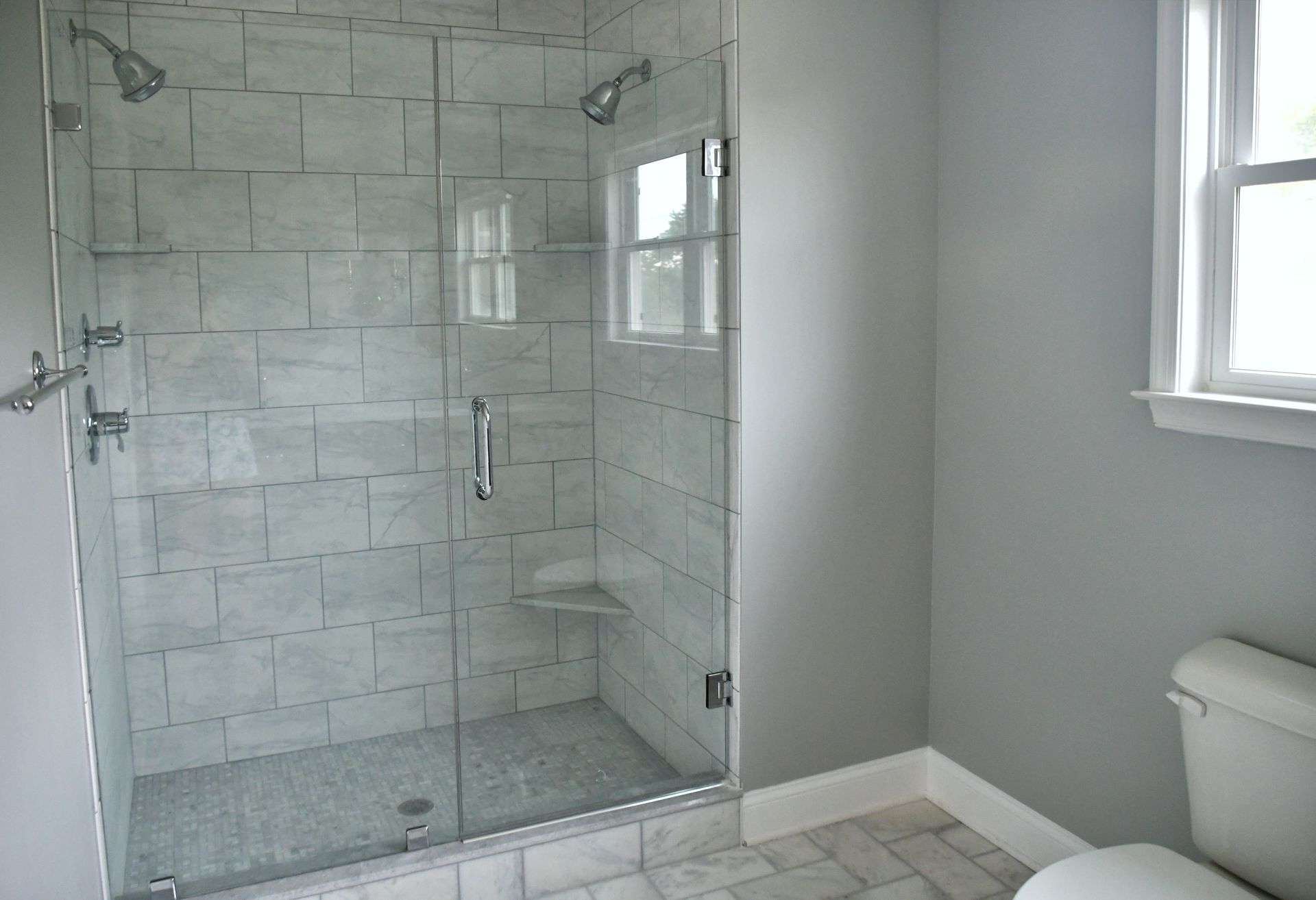 Empty tiled bathroom with a glass walk-in shower and window beside a toilet.