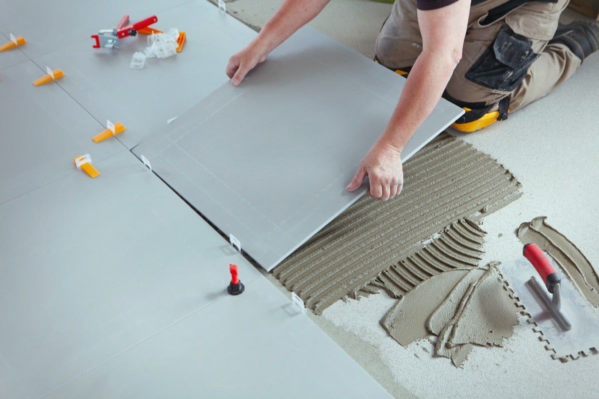 Person laying gray floor tile over adhesive with spacers and trowel on a subfloor