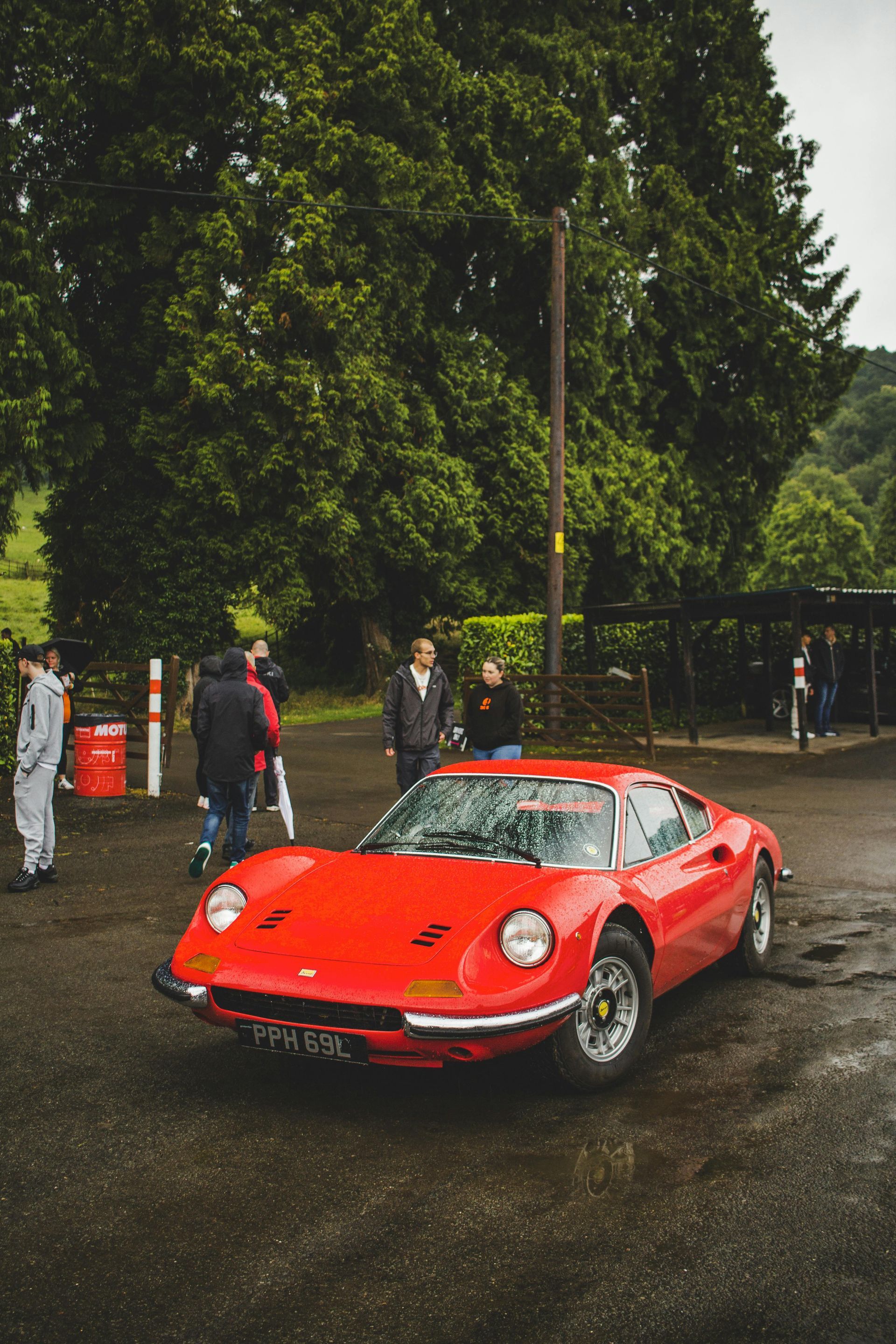 Voiture de sport Ferrari Dino rouge garée, personnes à proximité, chaussée mouillée, arbres verts.