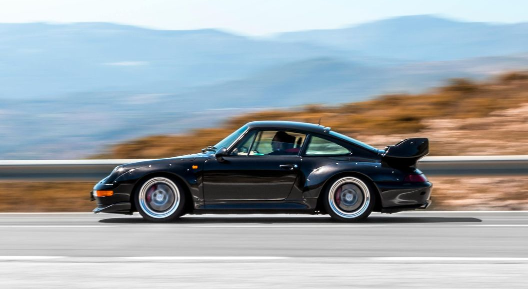 Une Porsche 911 Turbo noire file à toute allure sur une route avec des montagnes en arrière-plan.