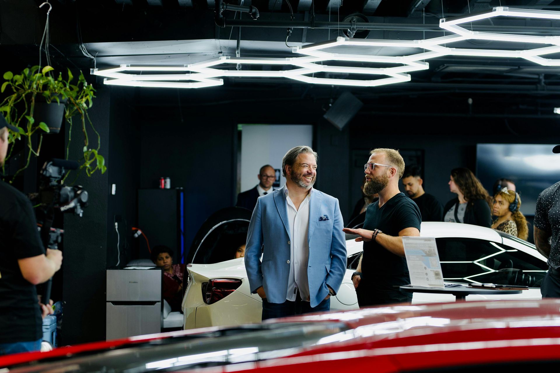 Des hommes en costume et en tenue décontractée discutent près d'une voiture moderne dans un showroom éclairé par des luminaires hexagonaux.