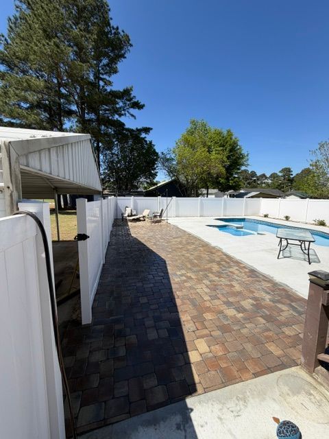Backyard patio with brick walkway, white fence, trees, and a small blue swimming pool under a clear sky
