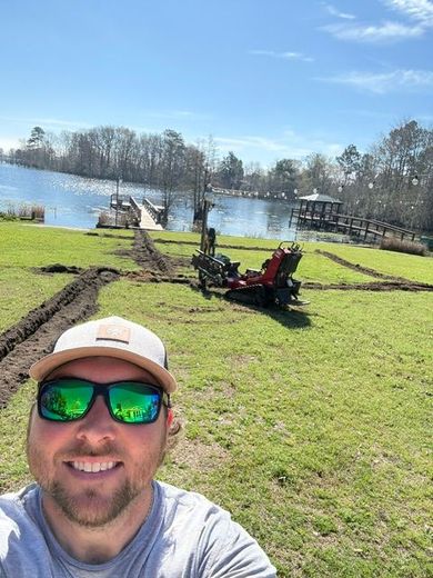 Man taking a selfie in a sunny yard by a lake, with a dock, trees, and a red lawn tractor in the background