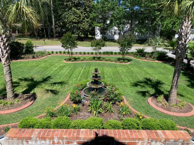 Landscaped garden with a small tiered fountain centered on a bright green lawn, framed by palm trees.