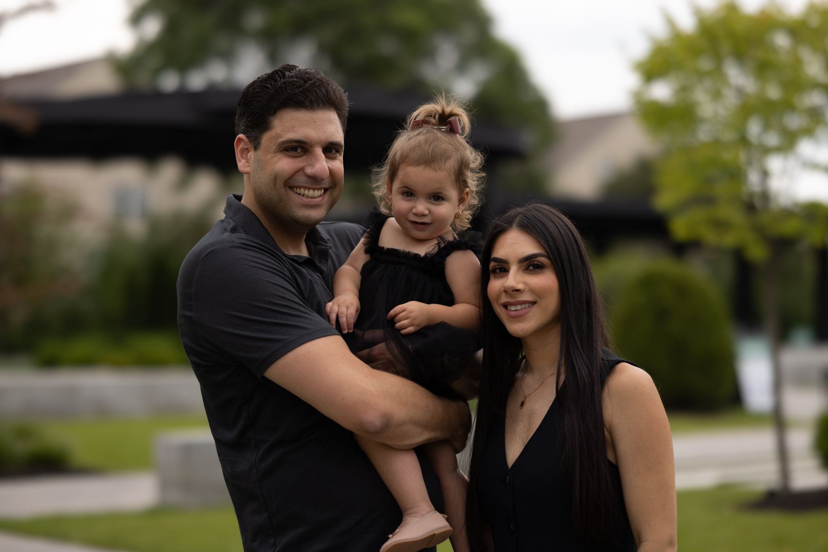 Family of three smiles outdoors: man holding child, woman stands beside them, all wear black.