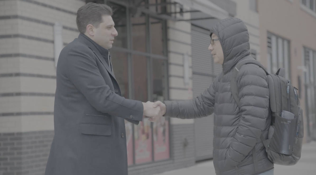 Two men shaking hands in front of a building. One wears a coat, the other a hooded jacket and backpack.