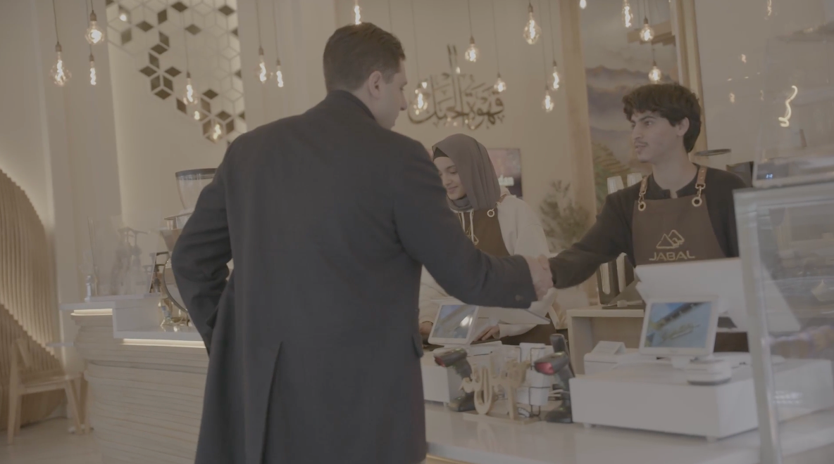 Man in coat at cafe counter, shaking hands with barista wearing apron.