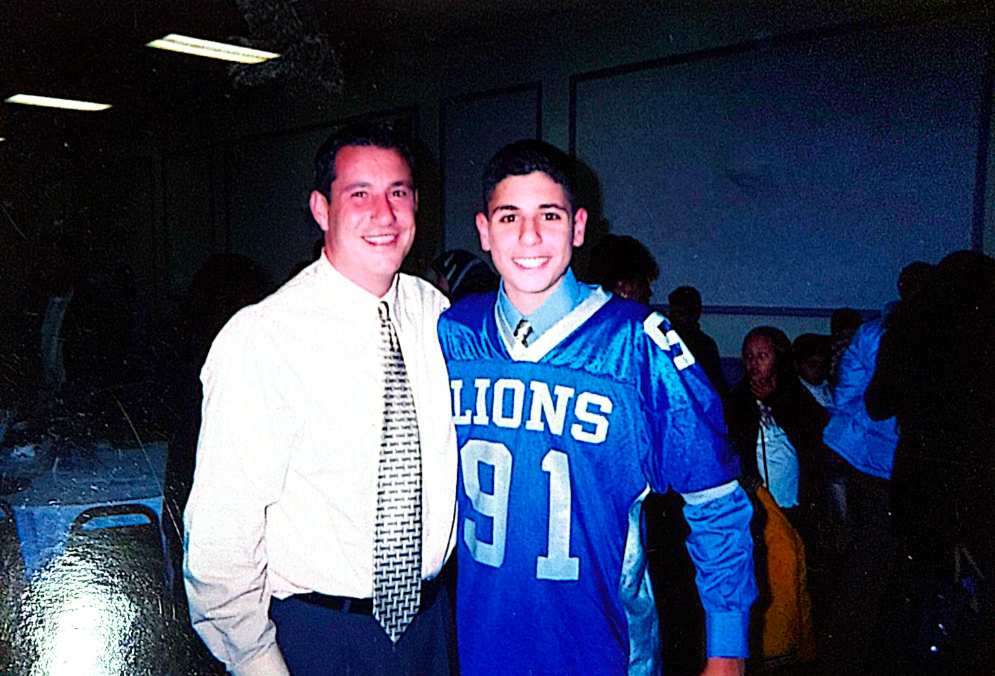 Man in button-up shirt and tie stands with a person in a blue football jersey.