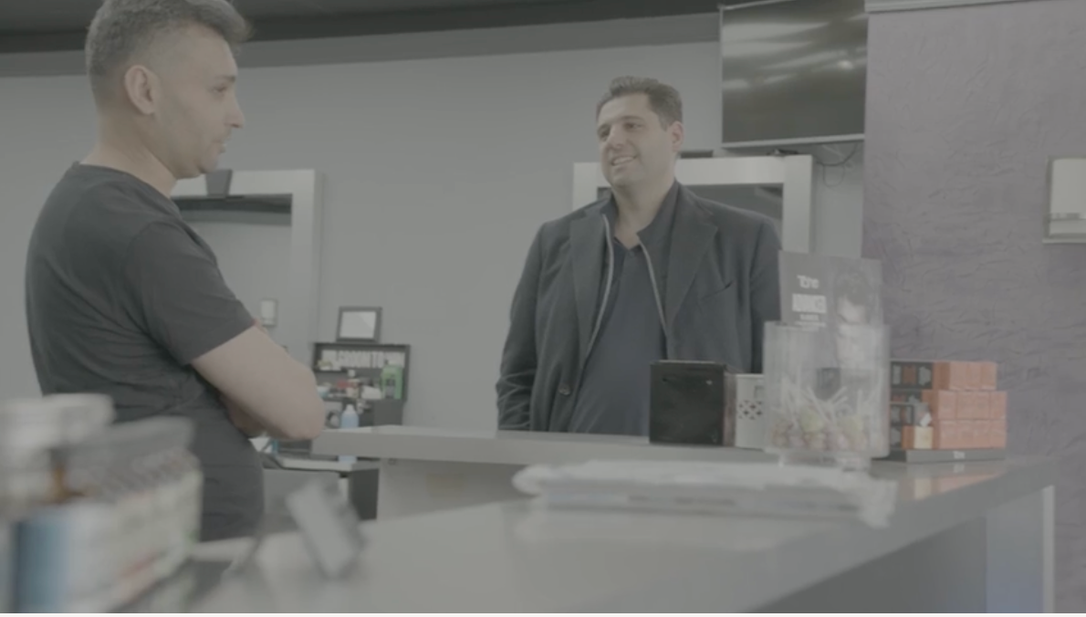 Two men talking at a counter in a business setting. One wears a black shirt, the other a black jacket.