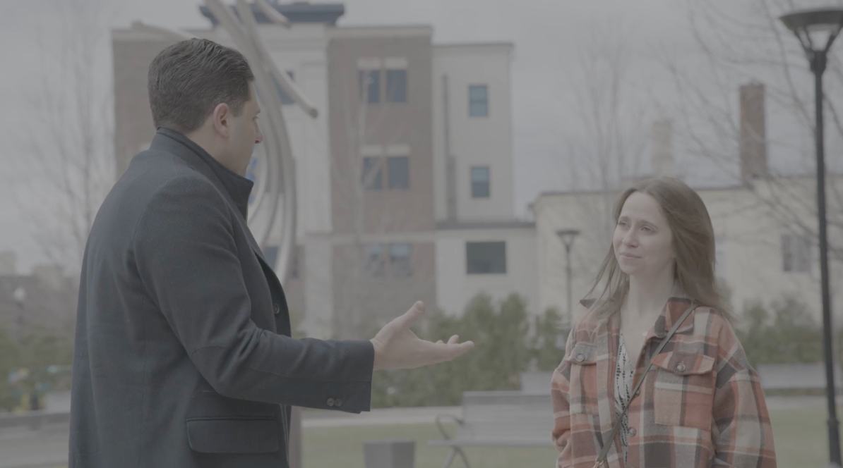 Man gesturing, talking to woman outdoors, overcast day. Buildings and trees in the background.