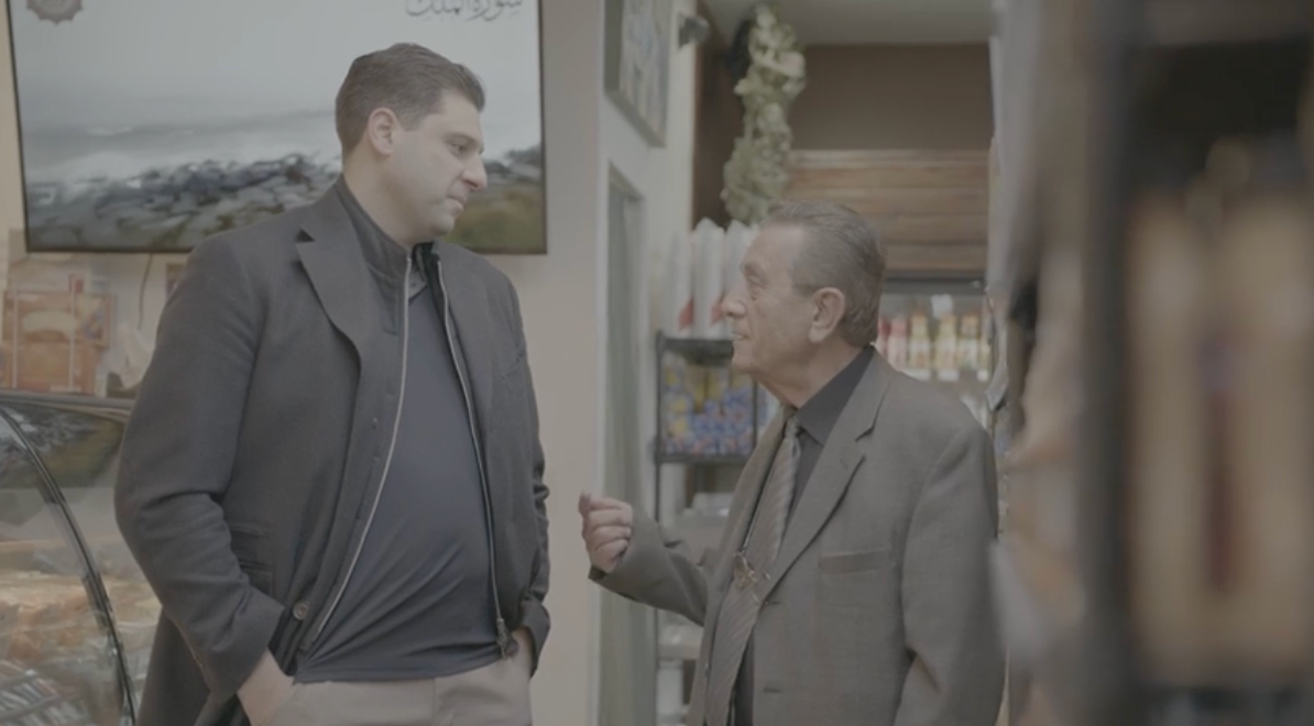 Two men talking inside a shop. One points while speaking, the other listens with hand in pocket. Shelves in background.