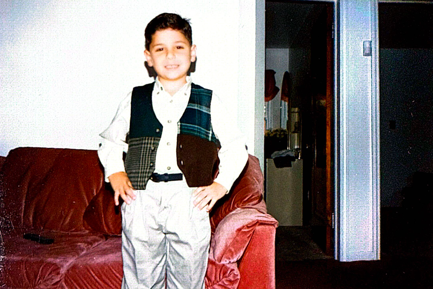 Young person in a vest and dress pants stands in front of a red couch.