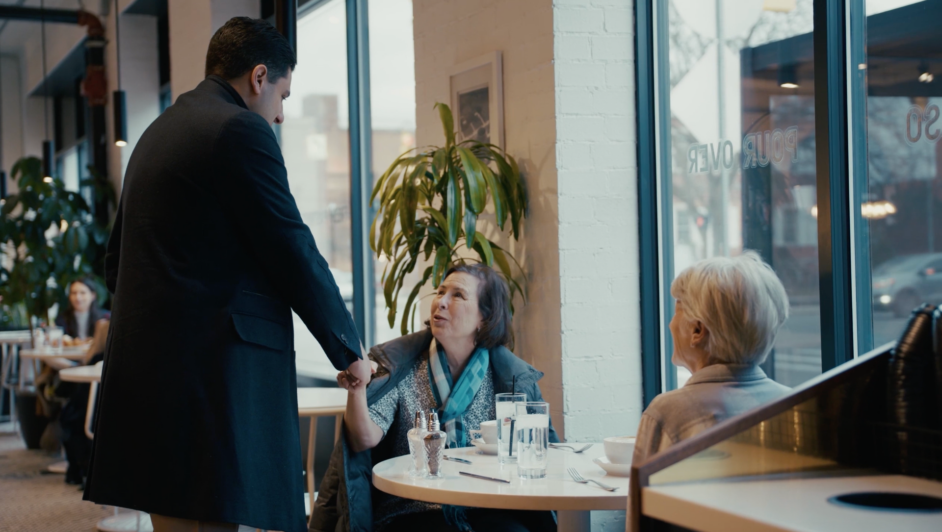 Man in coat greets two people at cafe table. Windows, plants, and other patrons are visible.