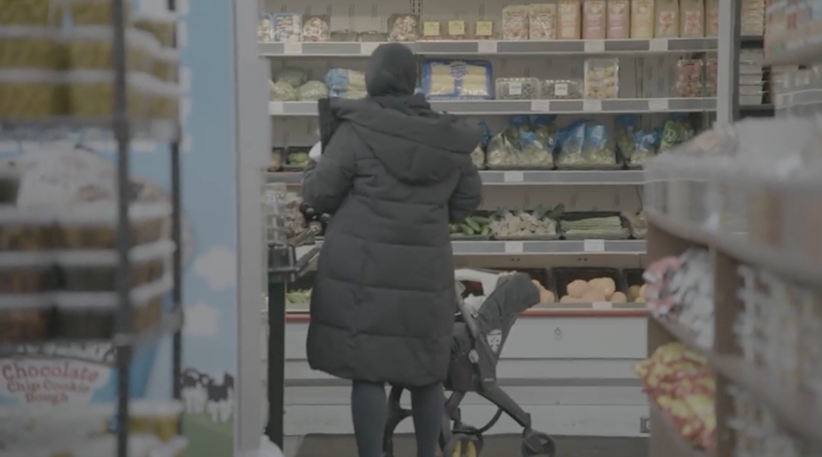 Person in dark coat shopping in a store, pushing a stroller; shelves with food visible.