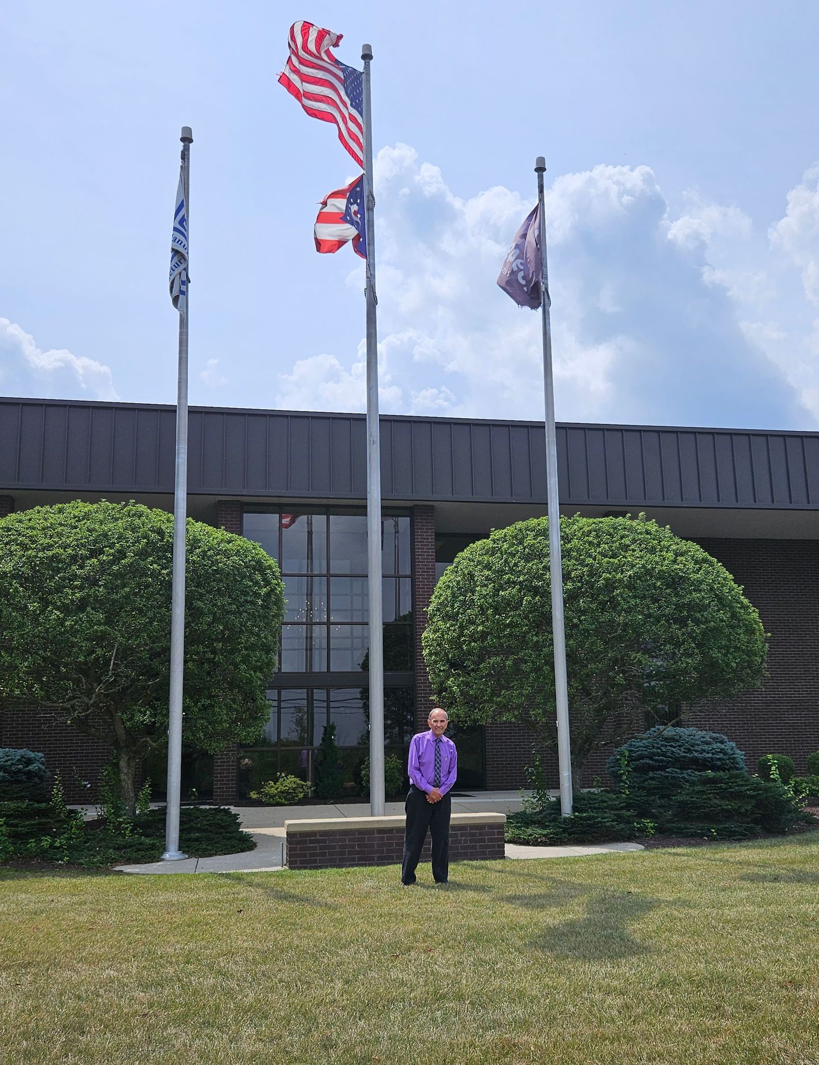 A man in a purple shirt stands in front of three flags