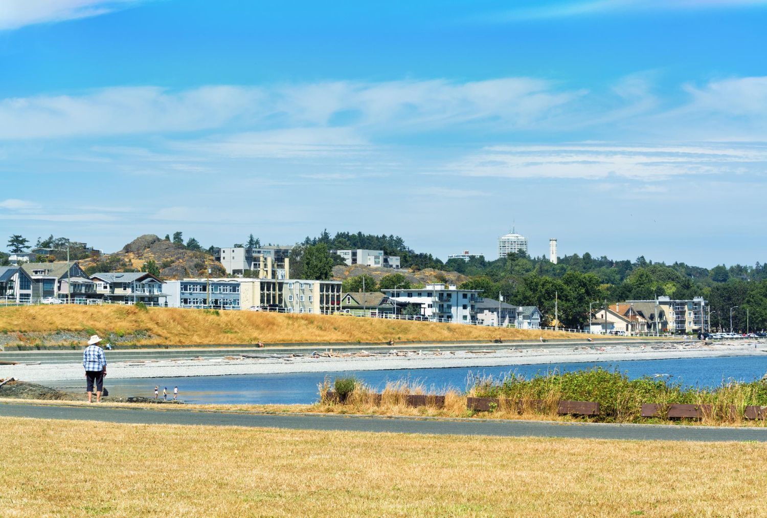 Person walks on a path near a beach, buildings in the background under a blue sky.