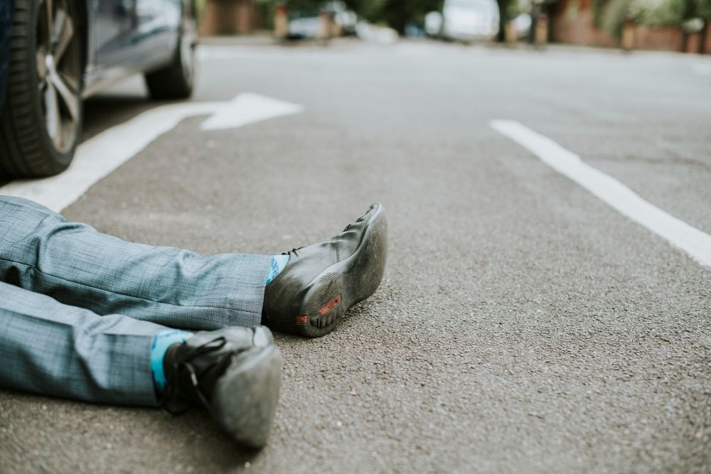 Man's legs on asphalt by car tire. He wears jeans, black shoes, on a street.