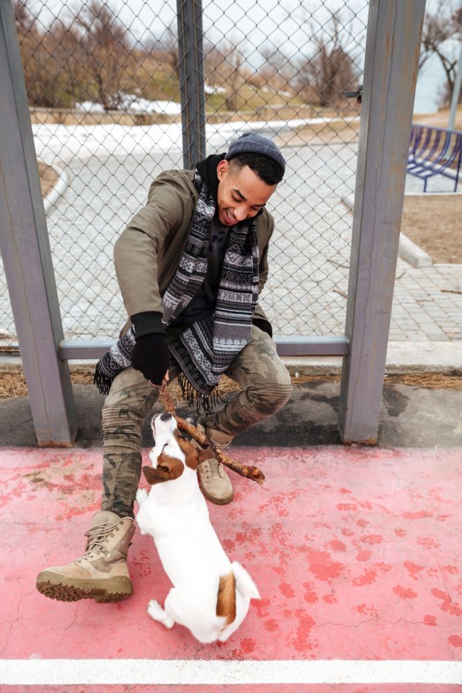 Person in green jacket and camouflage pants plays with a white and brown dog outdoors.