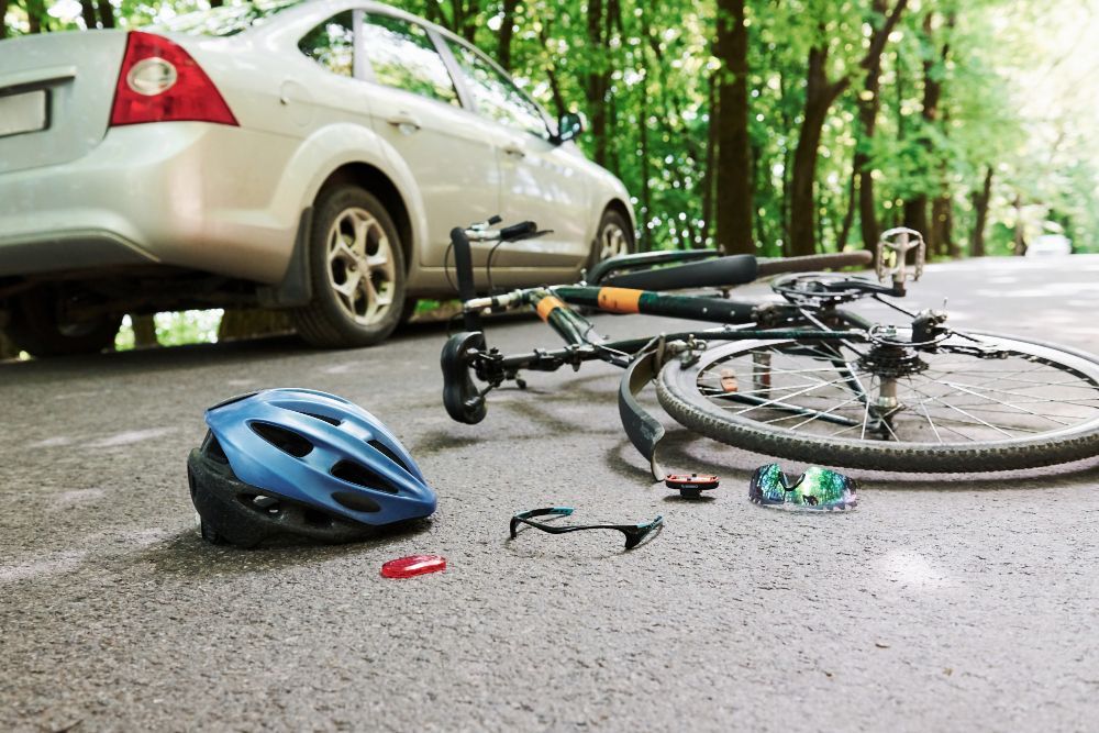 Car and bicycle collision scene: damaged bicycle on asphalt road near a silver car, helmet and debris scattered.
