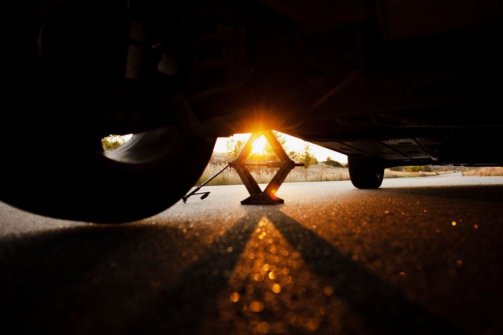 Car jack supporting a vehicle tire, sunlight behind it on asphalt.