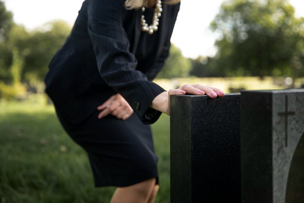 Woman in black suit at a cemetery, hand resting on a black headstone.