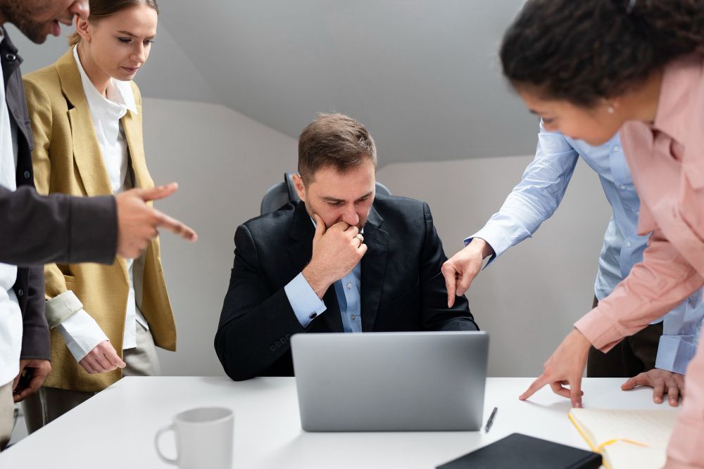 Business team surrounds a man at a laptop, pointing and looking concerned. Office setting with white table and neutral tones.