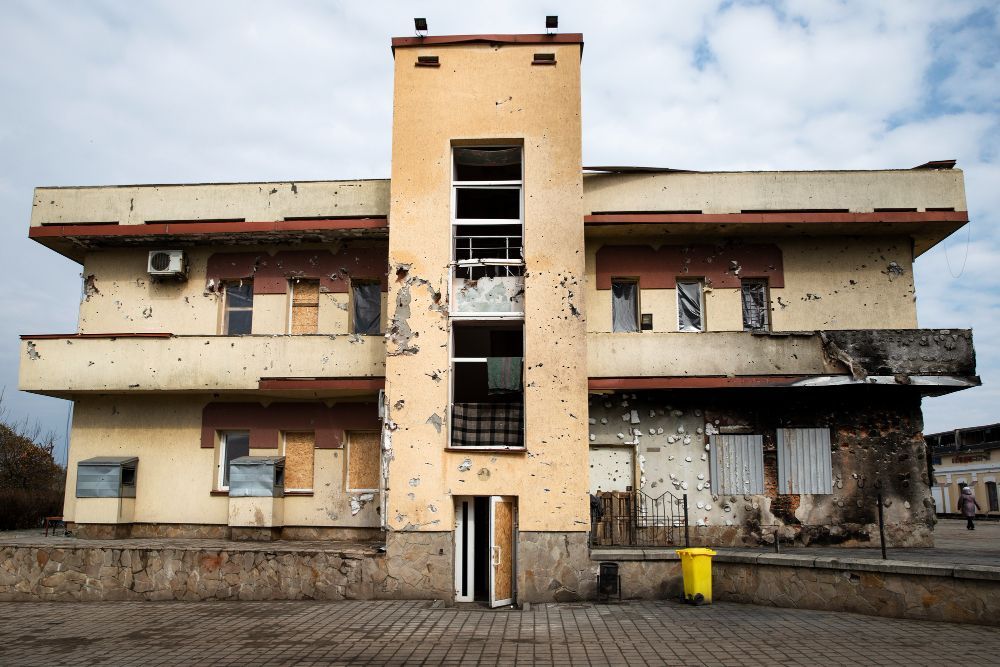 Damaged, two-story building with a central tower. Bullet holes, broken windows, and debris are visible. Exterior view.