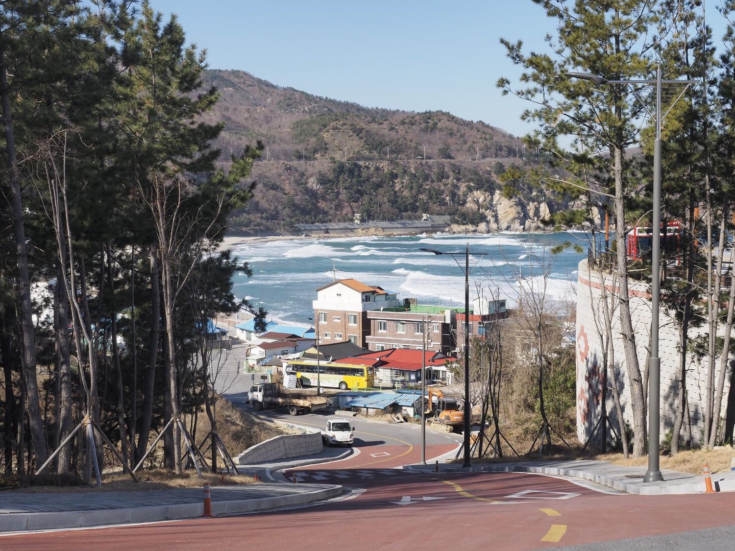 Coastal town with buildings nestled near a rocky shoreline and mountains, viewed through trees on a sunny day.